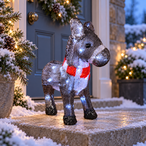 A glittery donkey-shaped outdoor light decoration with a red and white striped scarf stands on snowy stone steps in front of a decorated house door at dusk.