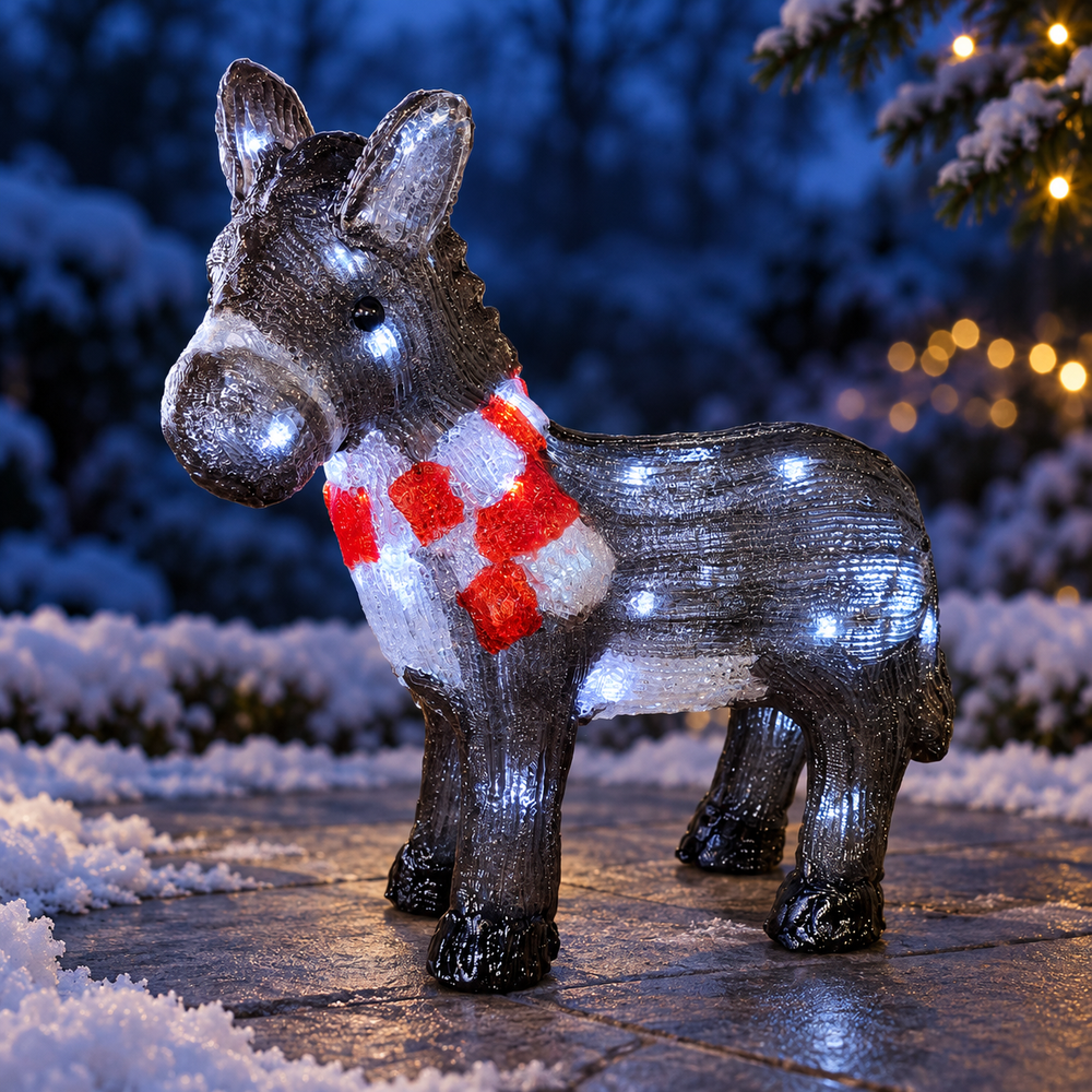 A decorative donkey figurine outdoors at night, covered in small white lights and wearing a red-and-white patterned scarf, standing on a stone surface with snowy ground and blurred holiday lights in the background.