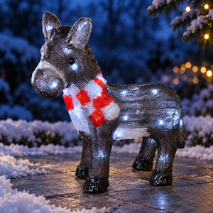A decorative donkey figurine outdoors at night, covered in small white lights and wearing a red-and-white patterned scarf, standing on a stone surface with snowy ground and blurred holiday lights in the background.