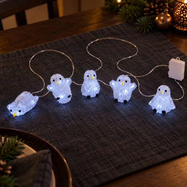 Five small penguin-shaped string lights glowing with a soft blue-white light, arranged on a dark textured placemat on a wooden table with holiday decorations in the background.