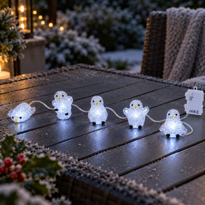 Five penguin-shaped outdoor string lights glowing on a dark wooden patio table, with a white power adapter visible on the right and a softly blurred garden background.