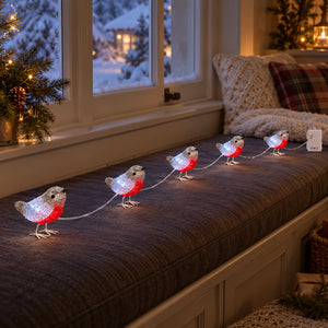 A string of decorative robin bird lights with glowing white bodies and red bellies arranged across a dark upholstered window seat in a cozy indoor setting.