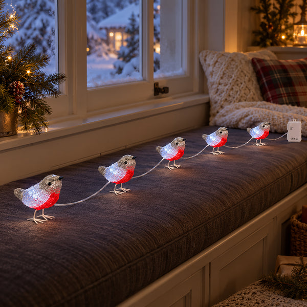 A string of decorative robin bird lights with glowing white bodies and red bellies arranged across a dark upholstered window seat in a cozy indoor setting.