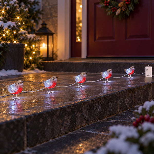 A string of holiday bird lights draped across wet stone steps in front of a decorated red door. Each bird has a silver glittery body and a bright red chest, with white strings connecting them to a white light box.