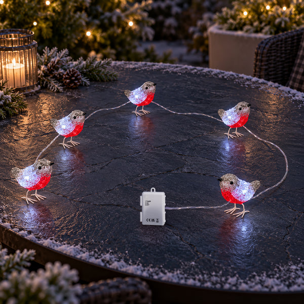 Outdoor Christmas string lights shaped like robins, with glittery silver bodies and bright red bellies, arranged in a circle on a dark stone patio. A small white power adapter is visible on the ground, and holiday greenery and warm lights appear blurred in the background.