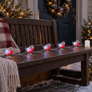 Outdoor Christmas string lights shaped like robins, with glowing clear bodies and bright red bellies, arranged in a line on a brown wooden bench. 