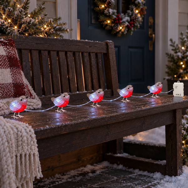 Outdoor Christmas string lights shaped like robins, with glowing clear bodies and bright red bellies, arranged in a line on a brown wooden bench. 