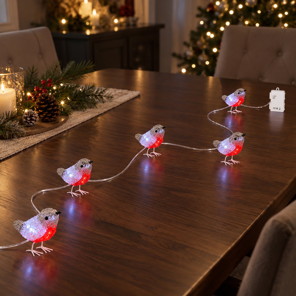 Lighted robin string lights shaped like small birds, glowing with red and white lights, arranged on a dark wooden dining table in a decorated holiday home setting.