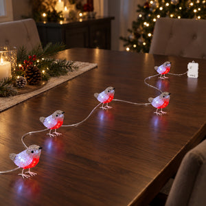 Lighted robin string lights shaped like small birds, glowing with red and white lights, arranged on a dark wooden dining table in a decorated holiday home setting.