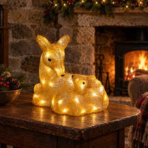 A golden glowing deer and baby deer figurine with warm white LED lights, displayed on a wooden table in front of a cozy stone fireplace with a burning fire.
