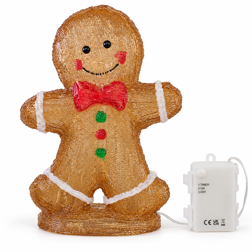 A lit gingerbread man holiday decoration standing on a stone surface outdoors at night, with snowy foliage and warm string lights in the background.