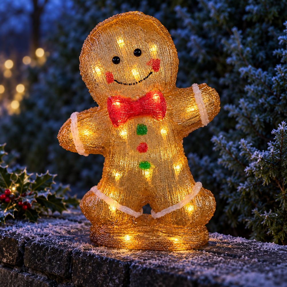A lit gingerbread man holiday decoration standing on a stone surface outdoors at night, with snowy foliage and warm string lights in the background.