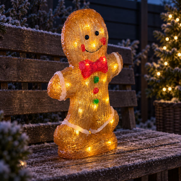 A glowing Christmas gingerbread man decoration with warm white LED lights, a red bow tie, and green and red buttons, displayed on a snowy wooden bench outdoors at night.