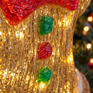 Close-up of a glittery gold Christmas stocking ornament with a red textured top and three vertical red and green button details, lit with warm golden lights.