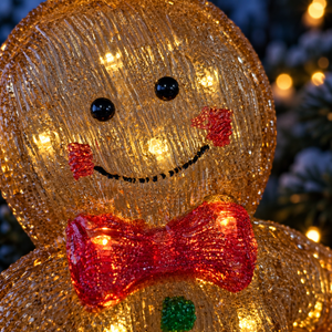 A close-up of a glowing Christmas gingerbread man decoration with a textured gold body, red bow tie, black eyes, and red cheeks, set against a blurred holiday background.