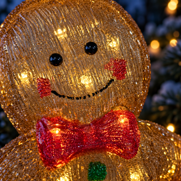 A close-up of a glowing Christmas gingerbread man decoration with a textured gold body, red bow tie, black eyes, and red cheeks, set against a blurred holiday background.