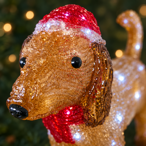 A close-up of a glittery dog Christmas ornament decorated with a red Santa hat and red scarf, set against a blurred Christmas tree background.