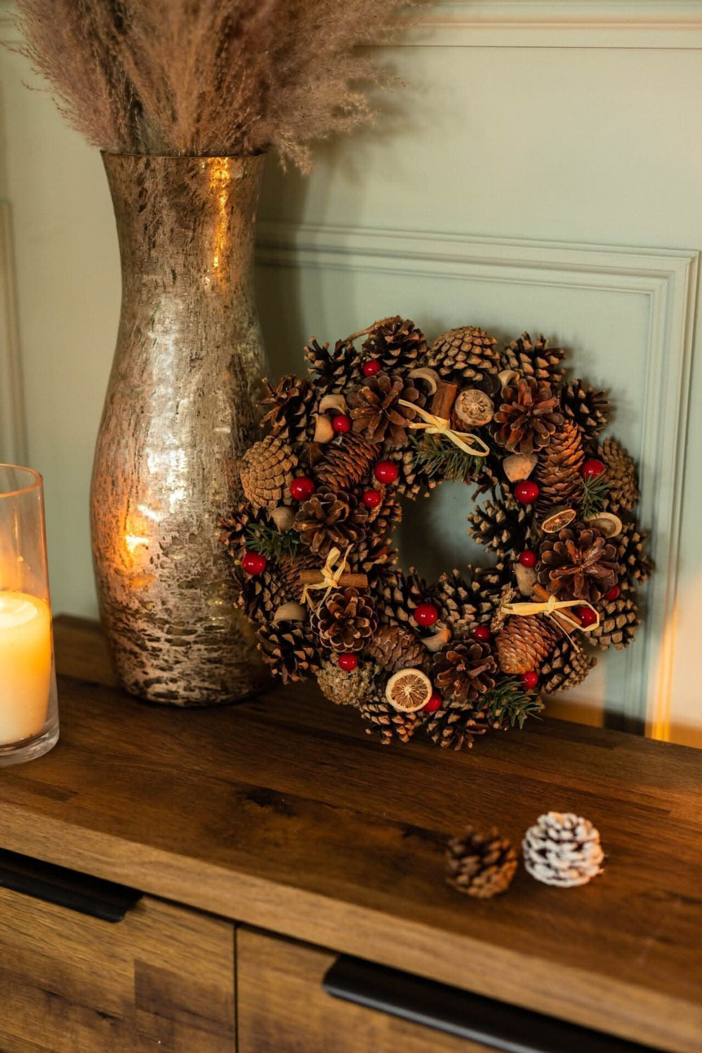 A luxury Christmas wreath made with dried fruits, pine cones, and berries, displayed on a wooden table next to a vase with dried flowers and candles.