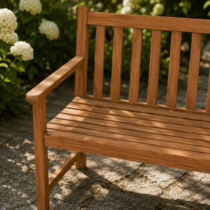 A close-up of a wooden garden bench with a warm brown finish, set outdoors on a stone patio with green foliage and white flowers in the background.