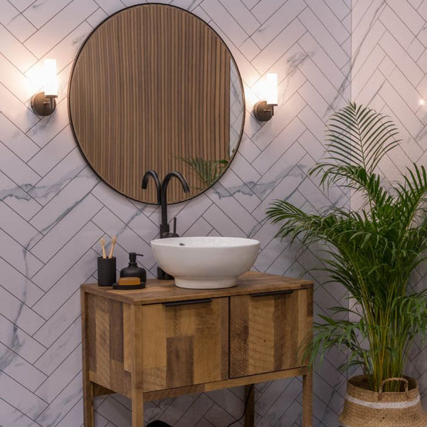 Bathroom with wooden vanity, round mirror, and plant against a tiled wall.