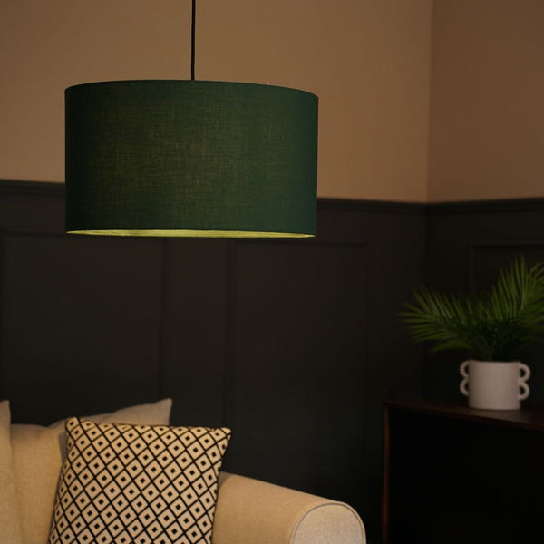 Dark green lampshade hanging above a beige sofa with a patterned pillow in a dimly lit room.