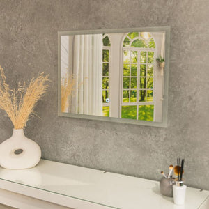 Mirror on a gray wall with a view of greenery, next to a vase with dried plants and a small table with items.