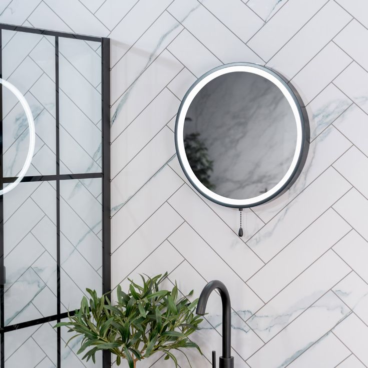 Round mirror with black frame on a marble wall with a plant and faucet in the foreground.