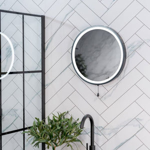 Round mirror with black frame on a marble wall with a plant and faucet in the foreground.