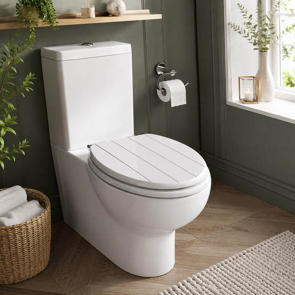 A white ceramic toilet with a slatted lid seat, shown installed in a styled bathroom with dark green walls, a window, plants, and a woven basket.
