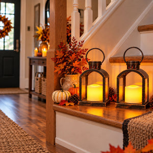 Two black metal lantern candles with warm yellow flames are displayed on a wooden staircase landing decorated for autumn, alongside pumpkins, pinecones, and fall foliage.