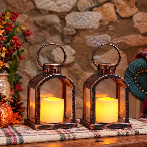 Two rustic bronze lanterns with glowing yellow LED candles, displayed on a plaid table runner against a stone wall background.
