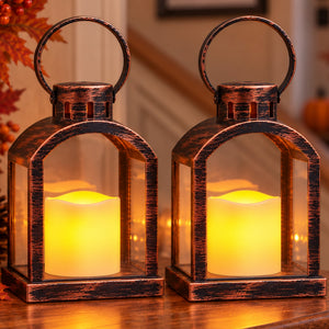 Two rustic dark bronze lanterns with clear glass panels and glowing yellow LED candles inside, displayed on a wooden surface in a warmly lit home setting.