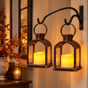 A double lantern wall sconce with two lit cream pillar candles, mounted on a beige wall in a warmly lit home setting.