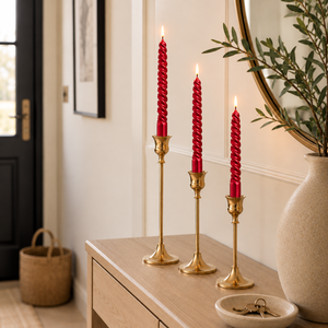 Three red spiral taper candles in gold candlesticks arranged on a light wood console table in a styled interior setting.