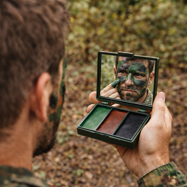 A man in camouflage clothing holds an open compact mirror in a forest setting, applying green face paint while looking into the mirror. The compact contains three makeup pans: green, brown, and black.