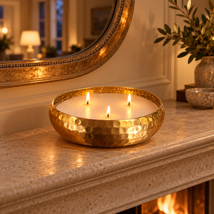 A hammered gold candle holder bowl containing three lit white candles, placed on a stone fireplace mantel in a warm, cozy interior setting with a mirror and plant in the background.