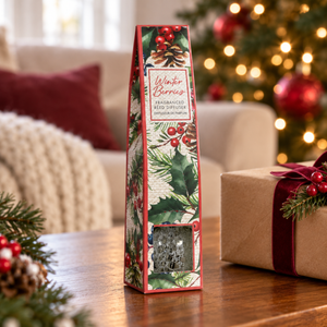 A tall, narrow decorative box of Winter Berries fragrance Reed Diffuser stands on a wooden table in a cozy holiday living room setting. The box features a festive red, green, and white holly and berry pattern, with a clear window near the bottom showing the textured reed diffuser inside.