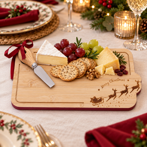 A rectangular bamboo holiday charcuterie board with a dark red border, engraved with 'Merry Christmas' and reindeer, styled on a table with cheese, crackers, grapes, and a cheese knife.