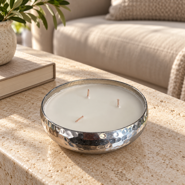 A hammered silver metal candle bowl filled with white wax and three wicks, placed on a beige stone table in a softly lit living room setting.