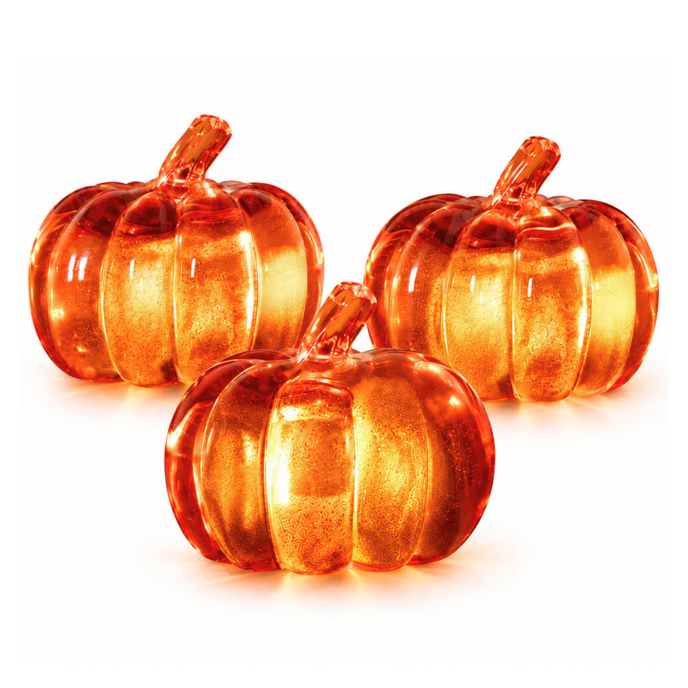 Three decorative glass pumpkins with warm orange and amber lighting, styled on a wooden table in a cozy living room setting with books, a candle, and fall leaves.