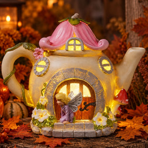 A decorative teapot-shaped fairy house ornament with a pink roof, glowing warm lights, a stone-textured base, and a small winged fairy figure in front of the door. The ornament is styled on a rustic surface with autumn leaves and pumpkins in the background.