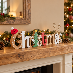 Wooden decorative letters spelling Christmas arranged on a wooden mantel with green garland, pinecones, and warm string lights in a cozy holiday setting.