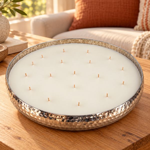 A large round candle with white wax and multiple wicks, set in a hammered silver metal tray on a wooden table in a cozy living room setting.