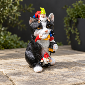 A black and white cat garden statue with three colorful garden gnome figures, displayed on a stone patio with blurred green plants in the background.