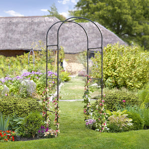 A black, powder coated steel garden arch standing in a garden bed, surrounded by greenery and flowers, with a thatched roof structure in the background.