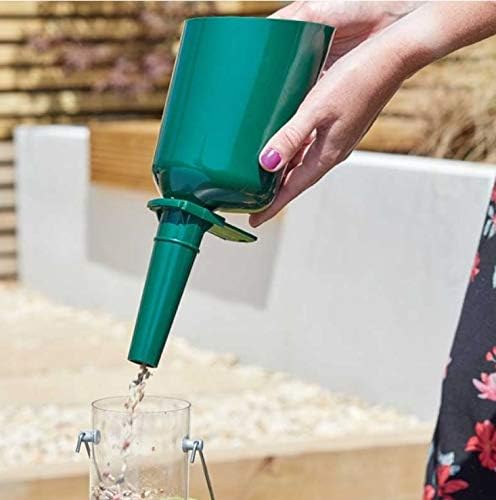 A hand with pink nail polish holds a green plastic funnel above a glass container, pouring a liquid. The background shows a blurred outdoor patio setting with a white planter and wooden fence.