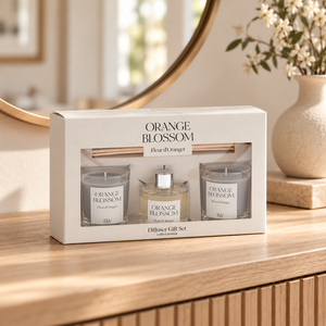 An orange blossom diffuser gift set in a white box, displayed on a wooden table in a warm, softly lit room. The set includes two glass jar candles labeled 'ORANGE BLOSSOM' and a central amber diffuser bottle with a silver cap, along with a wooden reed diffuser stick resting on top of the box.