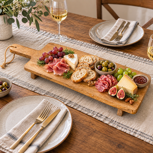 Raised mango wood cheese board with assorted cheeses, crackers, figs and chutney beside wine glasses.
