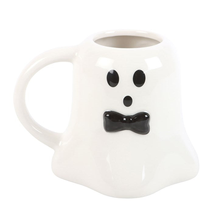 A white ceramic mug shaped like a ghost with a black face and bow tie, filled with coffee and styled on a cream knit blanket with cookies, pumpkins, a candle, and books in the background.