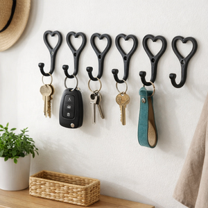 Six black heart-shaped wall hooks displayed on a white wall, holding keys, a car key fob, and a teal leather key fob strap. A woven basket, small potted plant, and a hat are partially visible in a styled home setting.